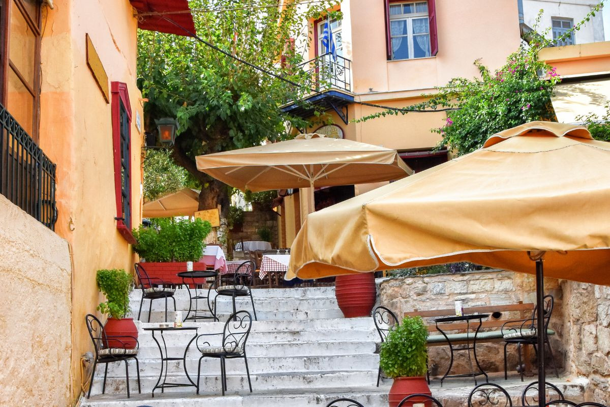 A cozy outdoor café with tables, chairs, and umbrellas on a stepped street, flanked by an orange wall on the left and a tree.